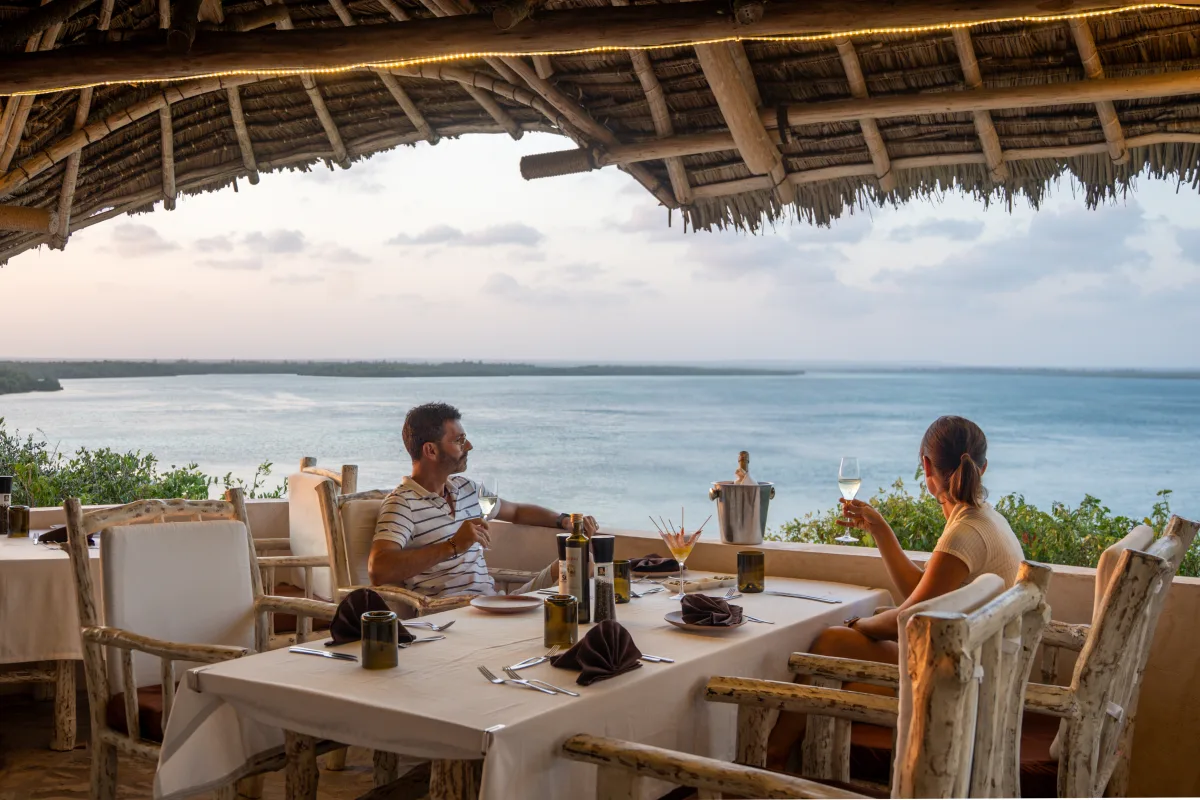Two guests raising a glass at a table under the makuti roof, the creek and the ocean stretching out behind them